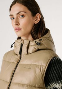 Beige gewatteerde vest met een hoge kraag en zwarte ritssluiting, met trekkoordverstellingen op de capuchon en een logo-detail aan de voorkant.