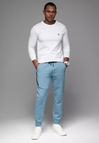 Męski strój składający się z białej koszuli z długim rękawem i jasnoniebieskich dresów z czarnymi akcentami, zestawiony z białymi sneakersami i zegarkiem.