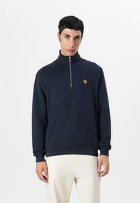 Mornarsko modra pulover z visokim ovratnikom, z zadrgo in majhnim zlatim emblemom. Tkanina deluje mehka z rebrastimi manžetami in robom.