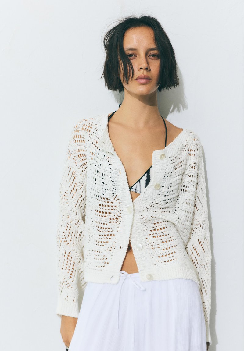 Jeune femme aux cheveux courts et foncés portant un cardigan blanc à maille ajourée sur un haut de bikini noir et un pantalon blanc ample, debout contre un mur blanc.