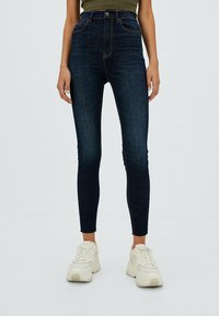 Jeans ajustados de cintura alta en denim azul oscuro con costuras naranjas. Presenta bolsillos funcionales y un dobladillo sin acabar. Combinados con zapatillas blancas.