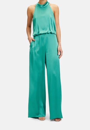 Femme portant une combinaison turquoise sans manches à col halter avec un pantalon à jambes larges et une main dans la poche, debout devant un fond uni.