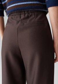 Bruine broek met een gladde textuur en een getailleerde pasvorm, voorzien van twee achterzakken en een elastische tailleband.