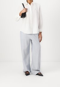 Bílá košile s dlouhým rukávem, světle modré široké kalhoty a černé špičaté boty. Košile má na hrudi malé logo.