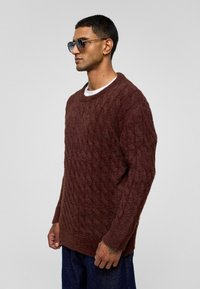 Bruine gebreide trui met een gestructureerd ruitpatroon, ronde hals en lange mouwen. Draagt over een wit shirt, gecombineerd met blauwe denim.
