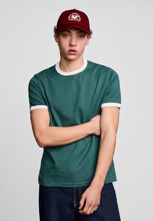 Jeune homme portant une casquette grenat avec un texte blanc, un t-shirt vert avec une bordure blanche et un jean en denim foncé, debout, un bras croisé.