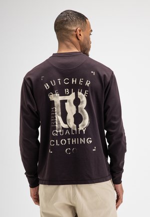 Mann, der von hinten zu sehen ist, trägt ein dunkles Langarmshirt mit dem Text ""Butcher of Blue Quality Clothing Co"" auf dem Rücken.