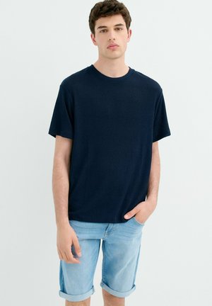 Joven con cabello corto y rizado, que lleva una camiseta azul marino de canalé y pantalones cortos de mezclilla azul claro con el dobladillo remangado, de pie contra un fondo liso y claro.