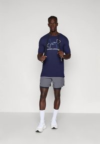 Jūrinės melsvos spalvos trumpomis rankovėmis marškiniai su Under Armour logo, derinami su pilkais šortais. Modelis laiko šokinėjimo virvę, vilkintis baltas sportines batus ir kojines.