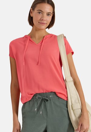 Jeune femme portant un top corail à manches courtes et un pantalon gris à cordon, portant un sac à épaule beige clair, debout devant un fond uni.