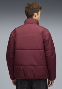 Veste matelassée bordeaux avec un col montant, un design matelassé et un ourlet élastique. Elle présente une texture lisse et est portée par une personne de dos.