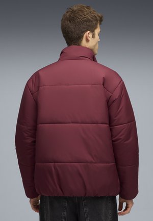 Burgundy pufferjakke med høj krave, quiltet design og elastisk kant. Den har en glat tekstur og bæres af en person set bagfra.
