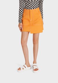 Gonna di jeans arancione con tasche frontali e orlo grezzo, abbinata a sandali platform bianchi. La parte superiore presenta un motivo a scacchi bianco e nero.
