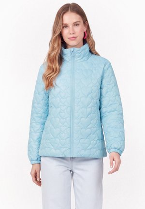 Jeune femme portant une veste matelassée bleu clair avec des motifs de cœurs, debout, les bras détendus, portant des boucles d'oreilles créoles roses et un jean clair.