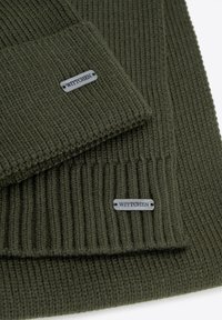 Ciemnozielony szal z prążkowanego dzianiny z teksturowanym wykończeniem, ozdobiony metalową metką z logo. Szal ma złożony brzeg, co zapewnia schludny wygląd.