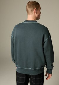 Tmavozelená mikina s okrúhlym výstrihom a voľným strihom. Má kontrastné prešitie a rebrované manžety. Zobrazené zozadu.