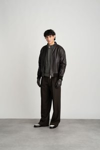 Μαύρο δερμάτινο μπουφάν bomber με ριγέ γιακά, γκρι πλεκτή μπλούζα από κάτω, φαρδύ καφέ παντελόνι, μαύρα γάντια και γυαλιστερά παπούτσια.