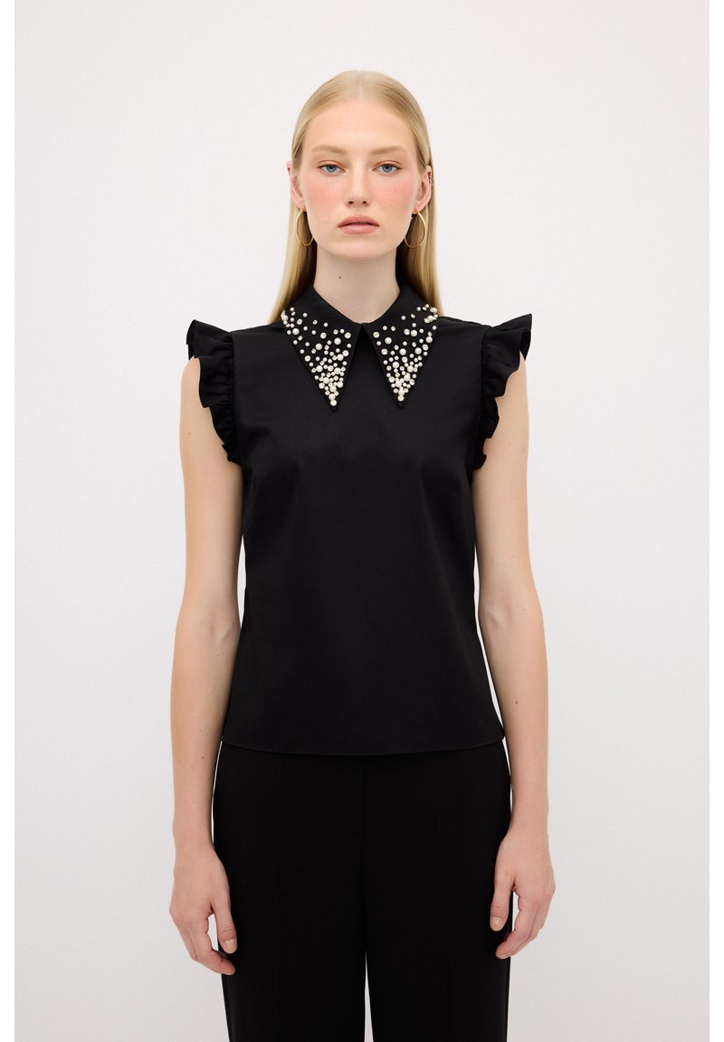 Sort ærmeløs bluse med frynse detaljer, der har perle-lignende udsmykninger på en spidse krave. Glat stof, tætsiddende design.