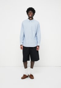Lichtblauw gestreepte overhemd met knopen en een zak, gecombineerd met zwarte knielange shorts, witte sokken en bruine loafers.