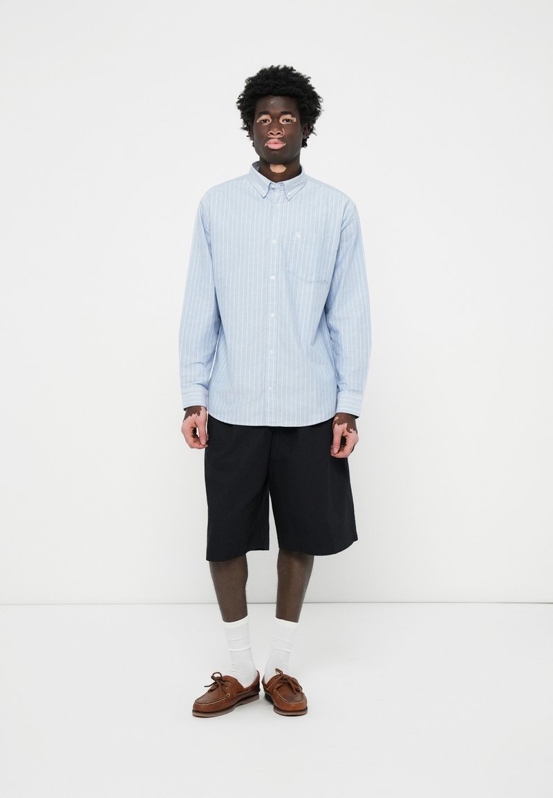 Chemise à rayures bleu clair avec une poche, associée à un short noir jusqu'aux genoux, des chaussettes blanches et des mocassins marron.