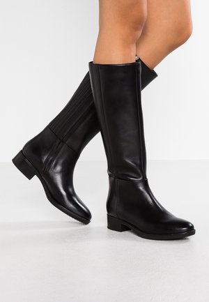 Jambes portant des bottes en cuir noires montantes jusqu'au genou avec des talons bas, debout et croisant légèrement un pied devant l'autre sur un sol blanc.