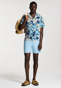 Kortärmad skjorta med knappar i blå och beige bladmönster, ljusblå shorts. Bär en vävd tote-väska. Bruna slidesandaler.