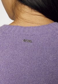 Pull violet tricoté avec un col rond côtelé et un logo argenté "BOSS" au dos. Texture douce avec une finition légèrement duveteuse.