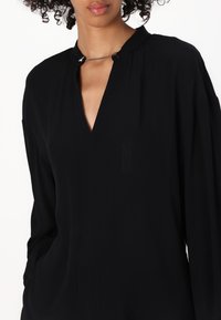 Blusa nera a manica lunga con scollatura a V e chiusura decorativa con catena al collo. Presenta un tessuto morbido con una caduta fluida e una leggera lucentezza.