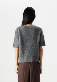 Maglione grigio a maniche corte realizzato in materiale fluffy. Presenta un collo rotondo, orlo a coste e una vestibilità ampia. Indossato con pantaloni marroni.