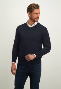 Marineblauwe v-hals trui van gebreide stof, met ribbelboorden en -zoom, gedragen over een wit overhemd met kraag.