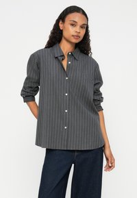 Camicia grigia con bottoni e strisce verticali bianche, maniche lunghe e taschino a sinistra sul petto. Realizzata in un tessuto liscio, presenta una vestibilità rilassata.