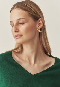 Auskaru pāris zelta tonī ar gludu, izliektu dizainu. Modelis valkā zaļu, spīdīgu V-veida kakla džemperi ar dziļu izgriezumu.