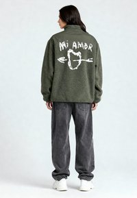 Veste polaire verte à col montant, arborant un motif graphique blanc comprenant "Mi Amor" et un cœur traversé d'une flèche. Portée avec un jean gris ample.