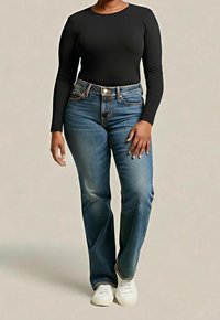 Personne portant un haut noir à manches longues, un jean taille haute bleu et des baskets blanches, debout devant un fond uni.