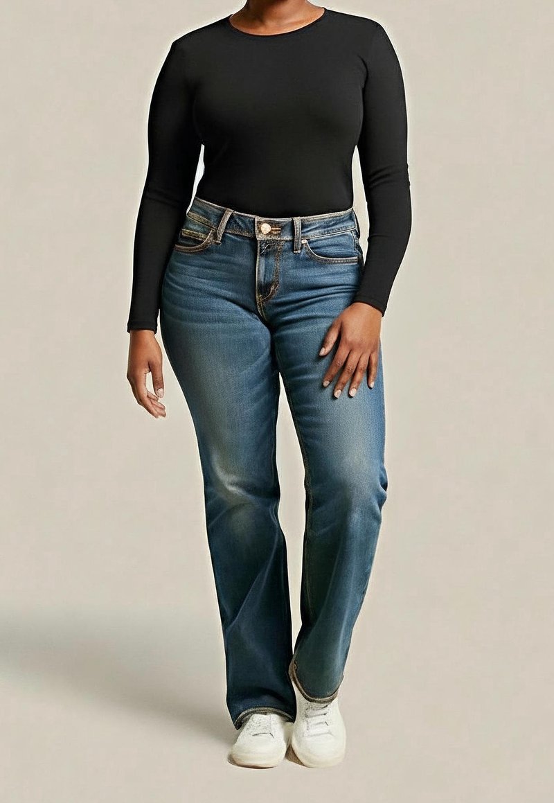 Personne portant un haut noir à manches longues, un jean taille haute bleu et des baskets blanches, debout devant un fond uni.