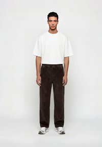 Άνδρας στέκεται με το βλέμμα προς τα εμπρός φορώντας λευκό πουκάμισο με κοντά μανίκια και χαλαρή εφαρμογή, σκουρό καφέ παντελόνι από κορδόνι και λευκά αθλητικά παπούτσια, μπροστά σε απλό φόντο.