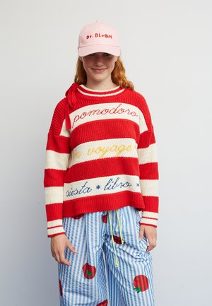 Πουλόβερ με κόκκινες και κρεμ ρίγες και μπλε και κίτρινο κείμενο· συνδυασμένο με παντελόνι με μπλε και λευκές ρίγες και κόκκινα σχέδια μήλων· ροζ καπέλο.