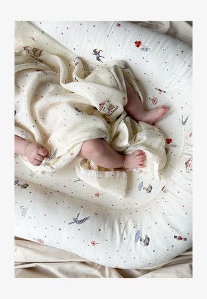 Bébé enveloppé dans une couverture crème avec des motifs sur le thème du cirque, allongé sur un coussin blanc orné de petits motifs d'étoiles et de personnages.
