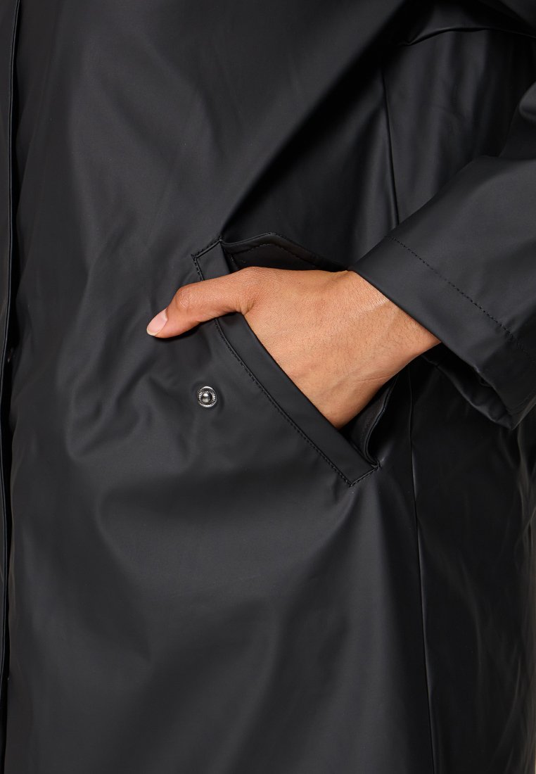 Main dans la poche d'un manteau noir imperméable avec un bouton-pression sur le rabat de la poche.