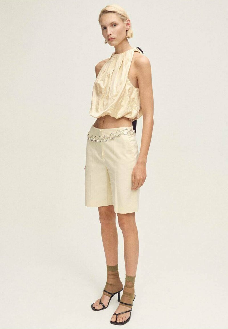 Cream-colored cropped top in shiny fabric, paired with beige shorts featuring a decorative belt. Model wears black heeled sandals with green socks.