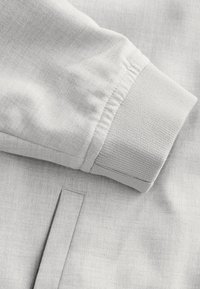 Jasnoszary rękaw kurtki z teksturowanym ściągaczem, gładkim materiałem z subtelnym wzorem oraz detalem szycia.