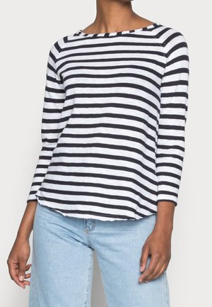 Persoon die een zwart-wit horizontaal gestreept shirt met lange mouwen draagt, gecombineerd met lichtblauwe spijkerbroek, staande tegen een effen achtergrond.