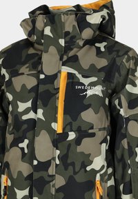 Kamouflagejacka i nyanser av grön och brunt, med orange detaljer, lager på lager fickor och en mjuk, texturerad yta. Varumärkeslogotypen är synlig.