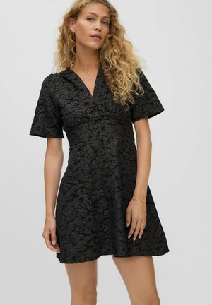 Woman with long curly blonde hair wearing a short black floral textured dress with short sleeves against a plain light background.