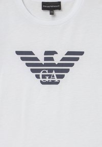 Bílý bavlněný triko s námořnickomodrou orlicí v logu, které má vodorovné pruhy a uprostřed silný text "GA". Klasický design s kulatým výstřihem.