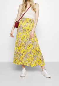 Falda maxi amarilla con un estampado paisley en rosa y verde. Combinada con una blusa beige y zapatillas blancas, complementada con un bolso rojo.