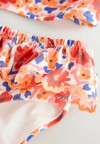 Színes virágmintás bikini szett fodros szélekkel, narancssárga, rózsaszín és kék virágokkal egy fehér alapon. Puha, rugalmas anyag.