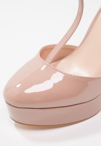 Chaussure à talon haut en cuir verni nude brillant, avec un bout arrondi, une semelle plateforme et une bride unique autour de la cheville. Texture lisse.
