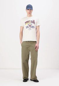 Jauns vīrietis, tērpies baltā "Tommy Jeans" grafiskajā T-kreklā, olīvzaļās biksēs, melnās kurpēs un zilos beisbola cepurē, stāv pret baltu fonu.