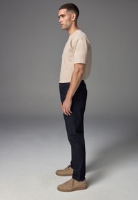 Jonge man staand in een beige t-shirt, donkere spijkerbroek en beige sneakers tegen een effen grijze achtergrond, gezicht naar links in profiel.